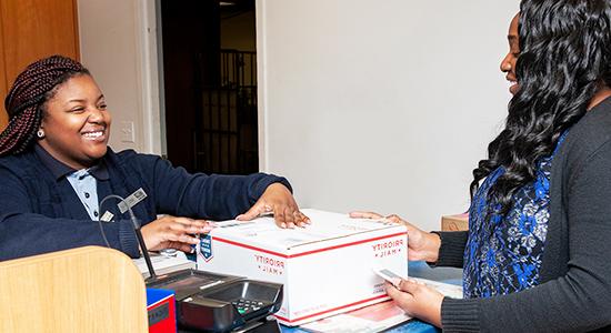 Person drops off Priority Mail package to USPS retail associate at a Post Office counter.